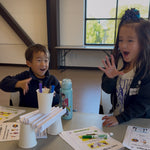 Two children sitting at a table indoors, smiling and gesturing while participating in a craft or activity session with paper cups, markers, and printed sheets on the table.